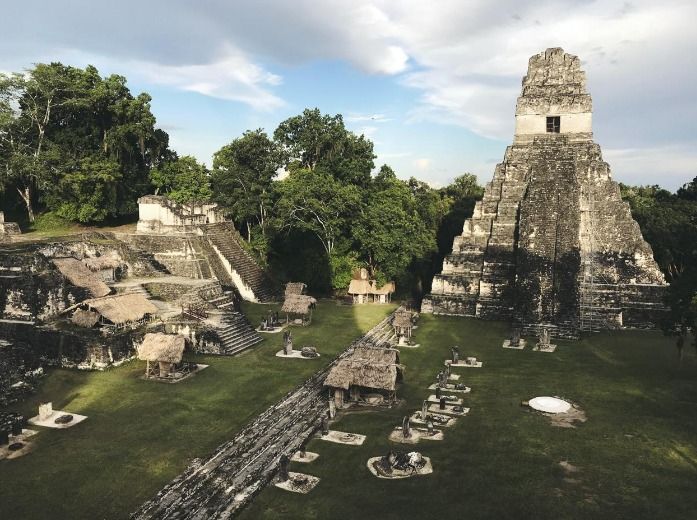 La luce del sole illumina un'antica piramide di pietra e rovine in un'ampia radura erbosa all'interno di una fitta giungla.
