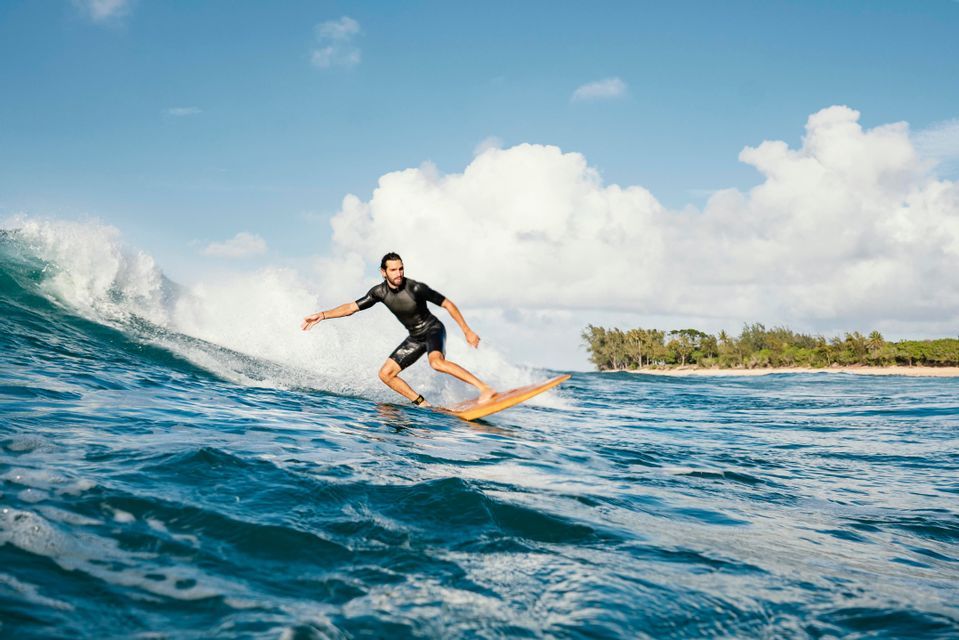 Un homme en combinaison de plongée noire surfe sur une vague bleue, avec une plage de sable visible à l'arrière-plan.