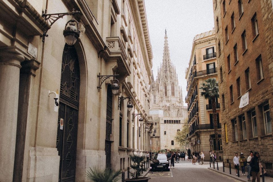 Una calle estrecha de la ciudad, flanqueada por edificios históricos de piedra, con la aguja de una catedral gótica visible a lo lejos.