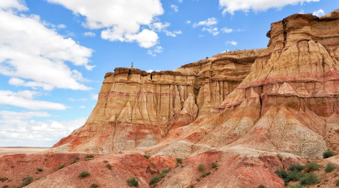 Une personne se tient au bord d'une formation rocheuse imposante, stratifiée, de couleurs rouge et jaune, sous un ciel bleu partiellement nuageux.
