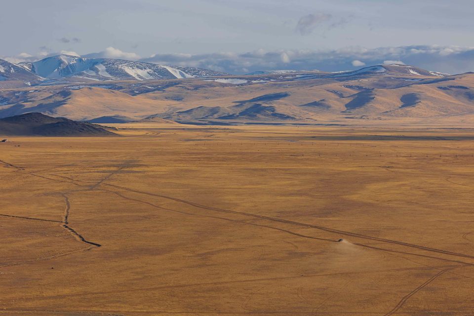 Un véhicule solitaire traverse une vaste steppe dorée, ses traces de pneus menant vers des montagnes enneigées au loin, sous un ciel nuageux.