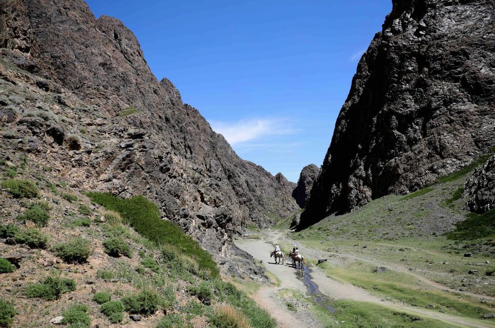 Deux personnes en voyage de groupe WeRoad montent à cheval à travers une vaste vallée montagneuse et rocheuse, près d'un ruisseau sous un ciel bleu.