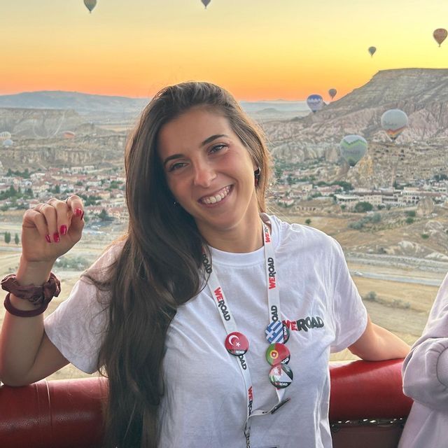 Una donna sorridente con una maglietta WeRoad posa in mongolfiera all'alba, con altre mongolfiere che fluttuano su un paesaggio roccioso.