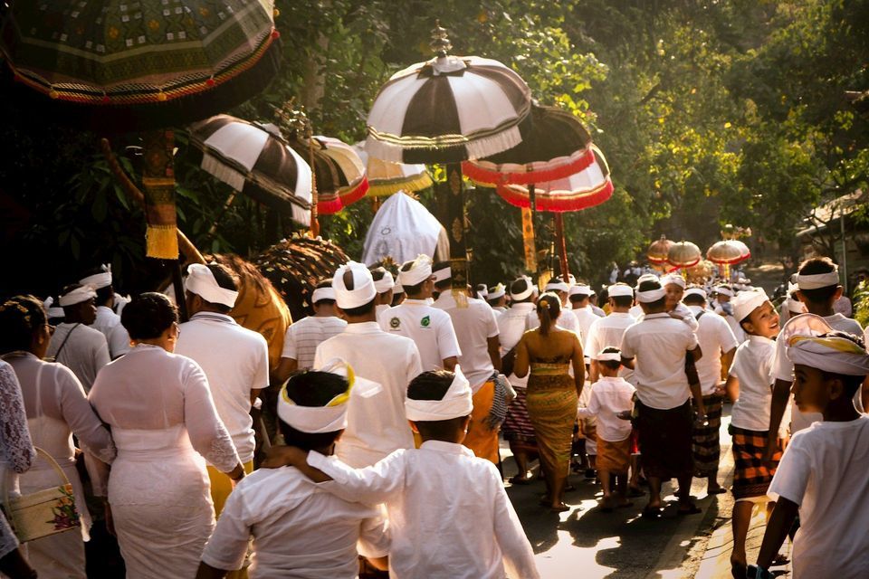 Un grande gruppo di persone in abiti tradizionali bianchi sfila in processione, portando grandi ombrelli cerimoniali lungo una strada alberata e illuminata dal sole.