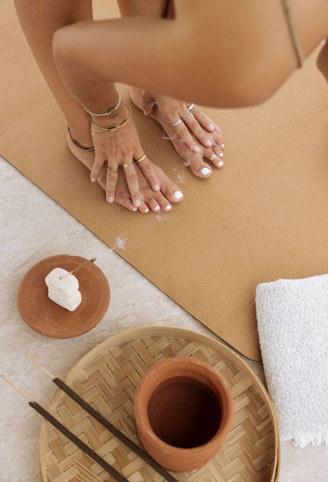 Una persona in posizione yoga di piegamento in avanti tocca i piedi su un tappetino marrone, con incenso che brucia e un asciugamano in primo piano.