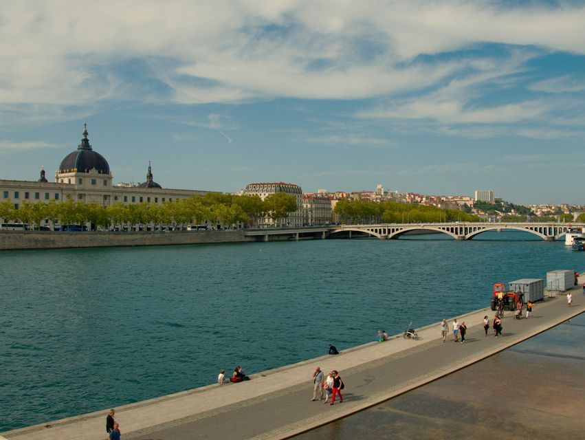 La gente passeggia e si rilassa su un lungofiume in una città, con un edificio a cupola e un grande ponte sullo sfondo.
