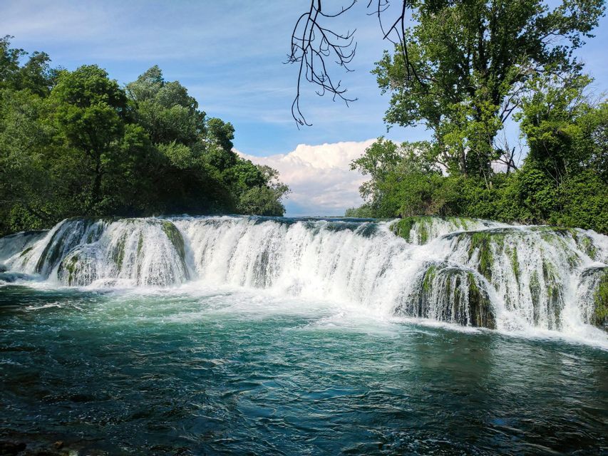 Une grande cascade s'écoule sur des rochers moussus dans une rivière turquoise, entourée d'arbres luxuriants.