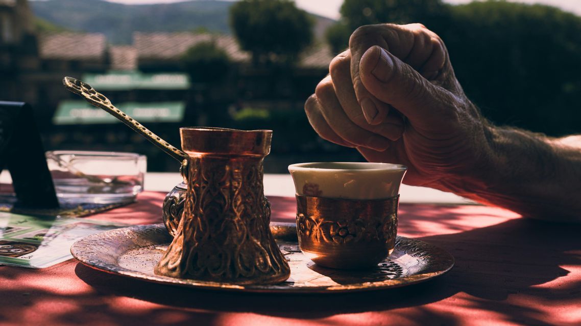 Une cafetière et une tasse traditionnelles en cuivre reposent sur un plateau gravé sur une table ensoleillée à côté d'une main.