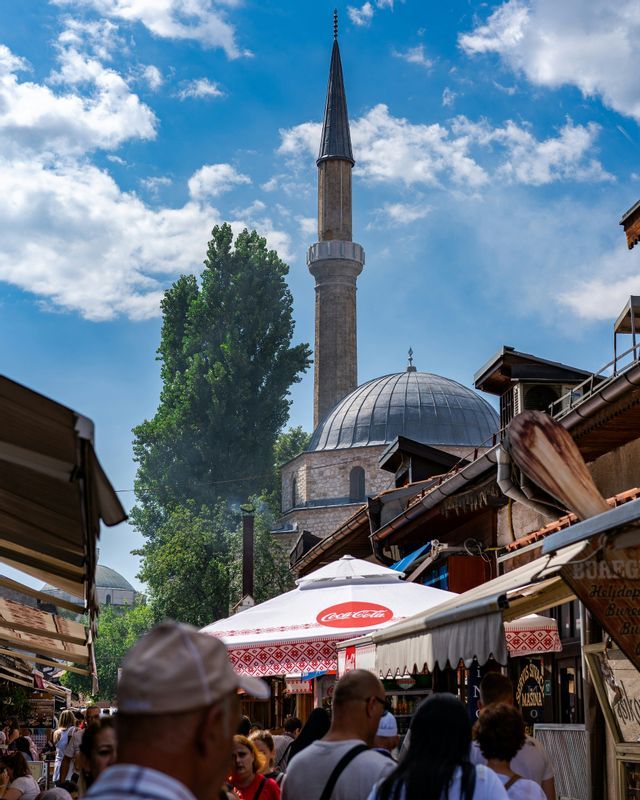 Une foule traverse une rue de marché animée, avec une mosquée en pierre et un minaret se dressant sous un ciel bleu nuageux.