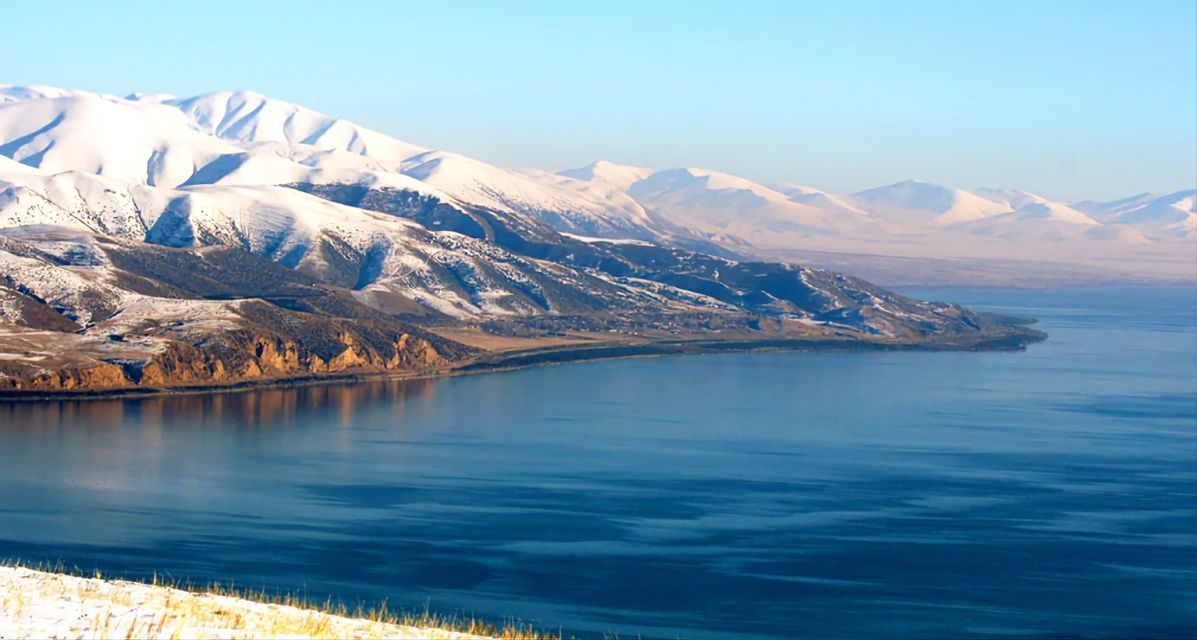 Schneebedeckte Berge treffen am Ufer eines ruhigen, tiefblauen Sees unter klarem Himmel.