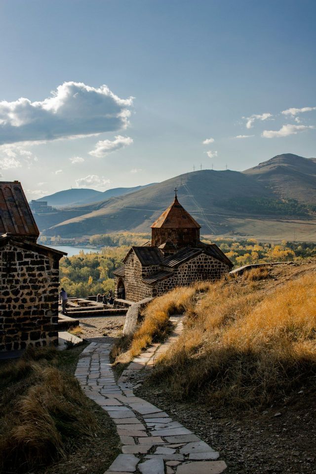 Ein Steinpfad schlängelt sich einen grasbewachsenen Hügel hinauf zu einer alten Steinkirche, die einen See und Berge im Hintergrund überblickt.