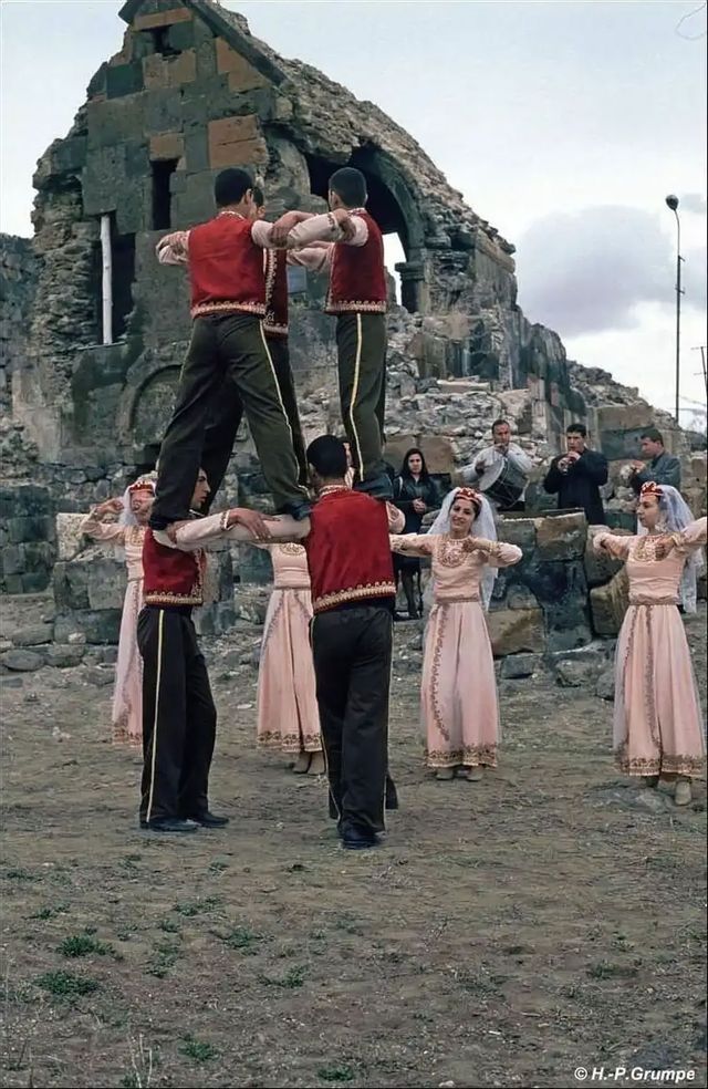 Eine Gruppe von Personen in traditioneller Kleidung führt vor steinernen Ruinen einen Volkstanz auf, wobei zwei Männer auf den Schultern anderer stehen.