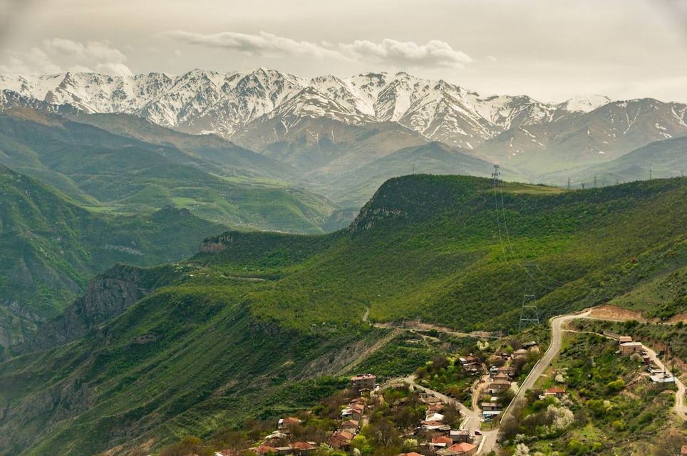 Ein kleines Dorf an einem üppig grünen Berghang mit gewundenen Straßen, vor der Kulisse hoch aufragender, schneebedeckter Berge.
