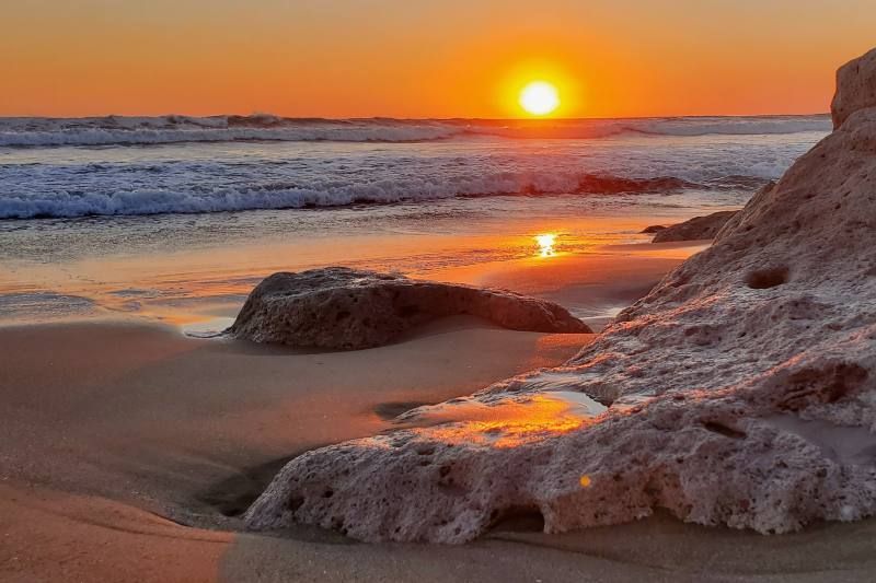 Il sole tramonta sull'oceano, proiettando un bagliore arancione sulle onde, sulla sabbia bagnata e sulle rocce sulla spiaggia.