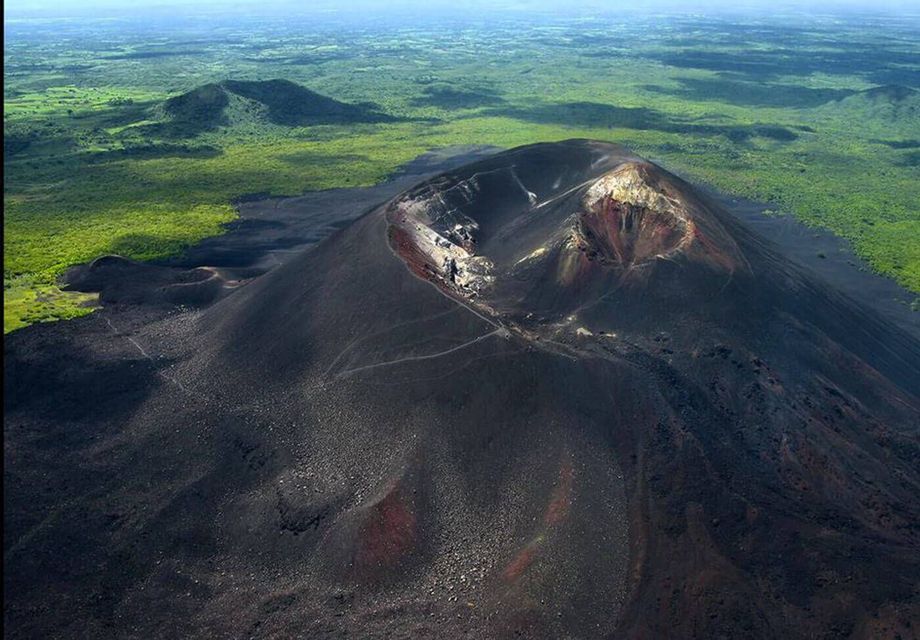 Una vista aerea di un vulcano scuro con escursionisti sul pendio che si dirigono verso il cratere, immerso in un lussureggiante paesaggio verde.
