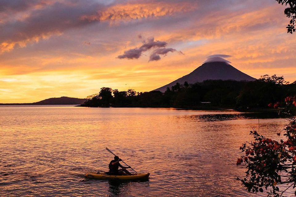 Una persona in kayak giallo su un lago al tramonto vibrante, con un grande vulcano stagliato sullo sfondo.