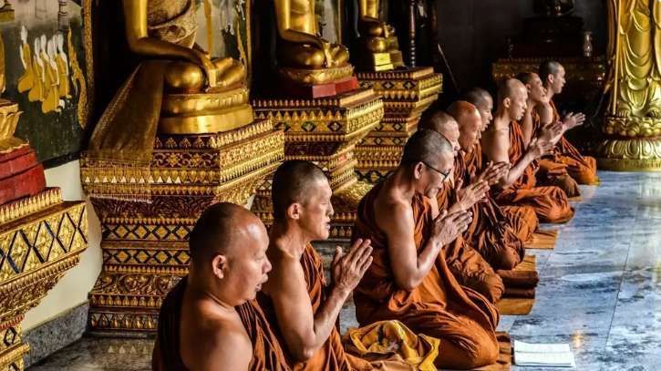 A line of Buddhist monks in orange robes kneeling in prayer inside a temple, in front of golden Buddha statues.