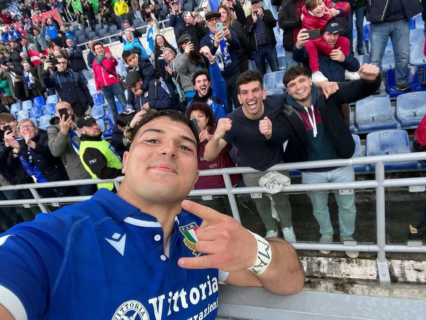 Un uomo in maglia sportiva blu si fa un selfie, facendo un gesto con la mano, con una folla di tifosi esultanti sugli spalti dello stadio alle sue spalle.