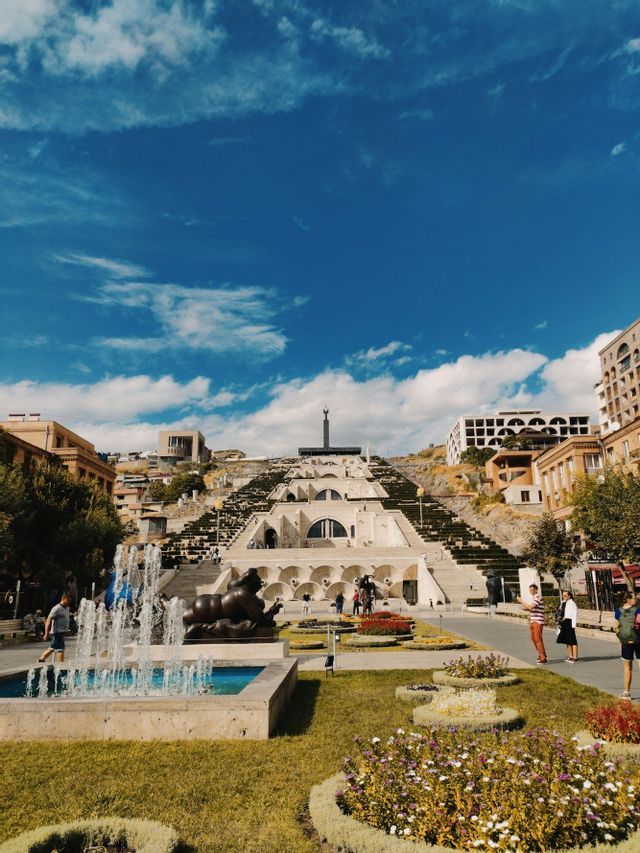 Eine monumentale terrassierte Steintreppe an einem Hang, gesehen von einem Park mit Springbrunnen, Skulpturen und Gärten unter blauem Himmel.