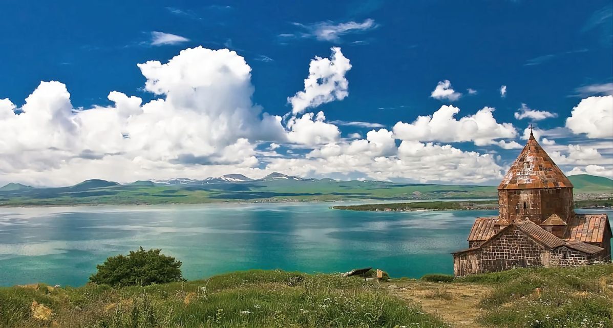 Eine alte Steinkirche mit kegelförmigem Dach steht auf einem grasbewachsenen Hügel mit Blick auf einen türkisfarbenen See und ferne schneebedeckte Berge.