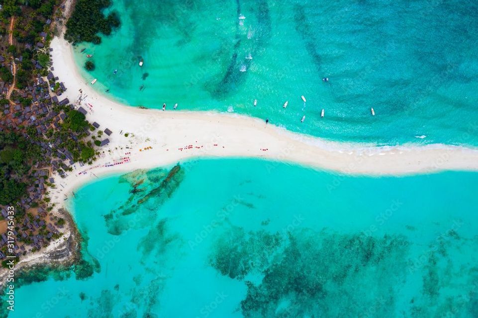 Vue aérienne d'un banc de sable blanc étroit s'étendant dans des eaux turquoises claires, avec de petits bateaux et un village côtier parmi les arbres.