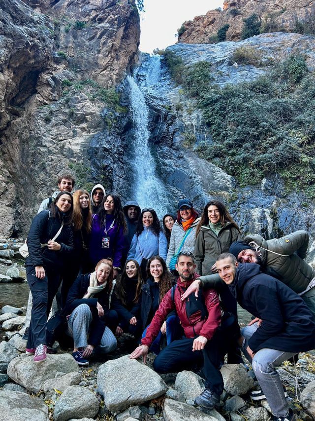 Un viaje en grupo de WeRoad posando para una foto en rocas en la base de una cascada que cae por un acantilado empinado.