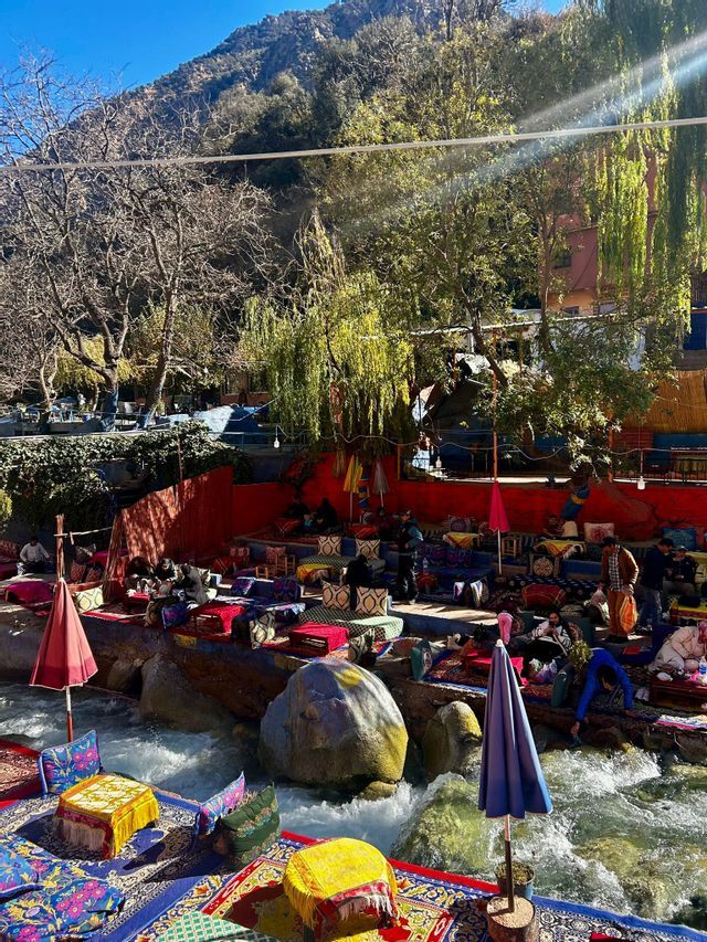 Un colorido café al aire libre con asientos sobre alfombras y cojines dispuestos en rocas, junto a un río caudaloso en un paraje montañoso y boscoso.
