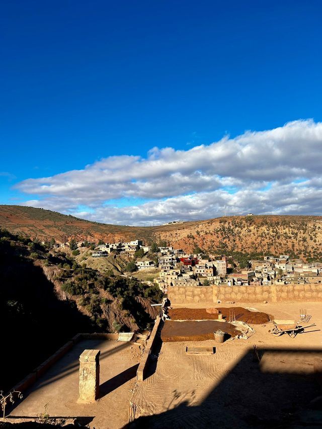 Una vista soleada desde una azotea, con vistas a un pueblo anidado en colinas áridas y rojizas bajo un cielo azul brillante y nubes.
