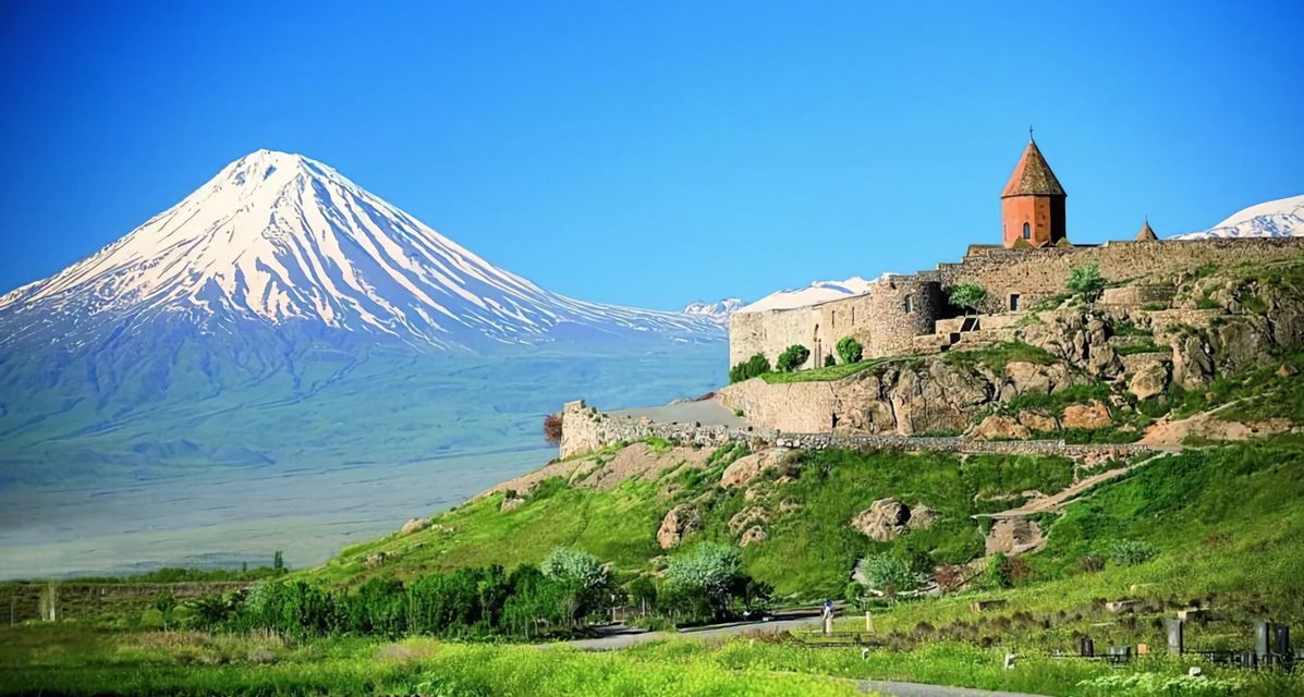 Ein Steinkloster auf einem grünen Hügel mit großem schneebedecktem Berg im Hintergrund unter klarem blauem Himmel.