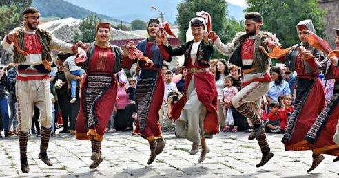 Eine Gruppe von Männern und Frauen in farbenfrohen Trachten tanzt im Freien einen Volkstanz vor Publikum und hält dabei Händchen.