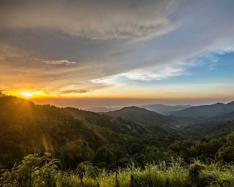 El sol se pone sobre un valle de montañas verdes y exuberantes, proyectando rayos de sol dorados a través de los árboles bajo un cielo parcialmente nublado.