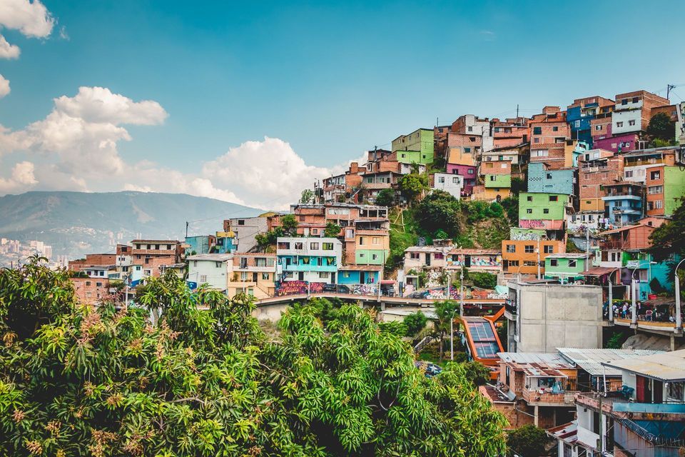 Un denso barrio de casas de colores cubre una colina verde, con una escalera mecánica exterior y montañas a lo lejos bajo un cielo azul.