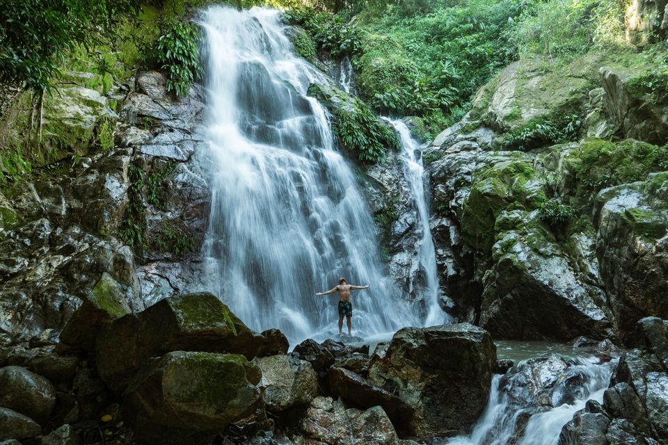 Ein Mann, oberkörperfrei und mit ausgestreckten Armen, sitzt auf den Felsen am Fuße eines Wasserfalls im Herzen eines dichten Dschungels.