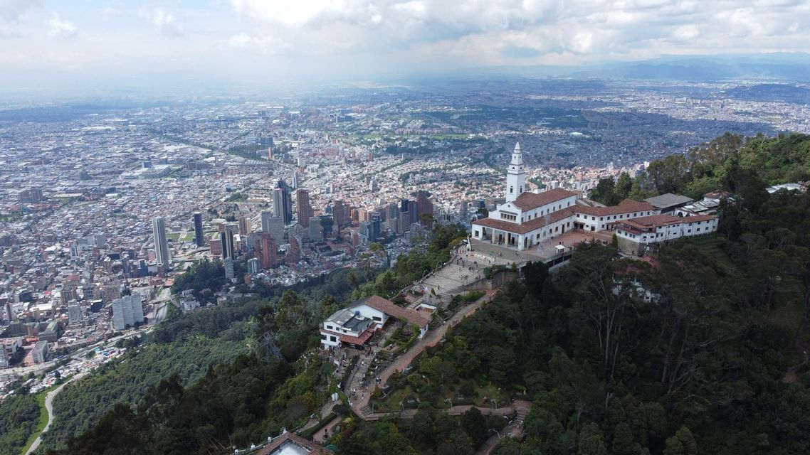 Vista aérea de un santuario blanco situado en una montaña boscosa, con vistas a una extensa ciudad moderna bajo un cielo nublado.