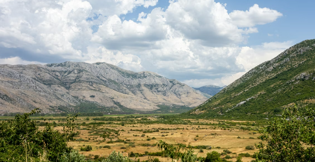 Un vaste paysage de vallée sèche, avec une montagne verdoyante d'un côté et une chaîne rocheuse de l'autre, sous un ciel bleu nuageux.