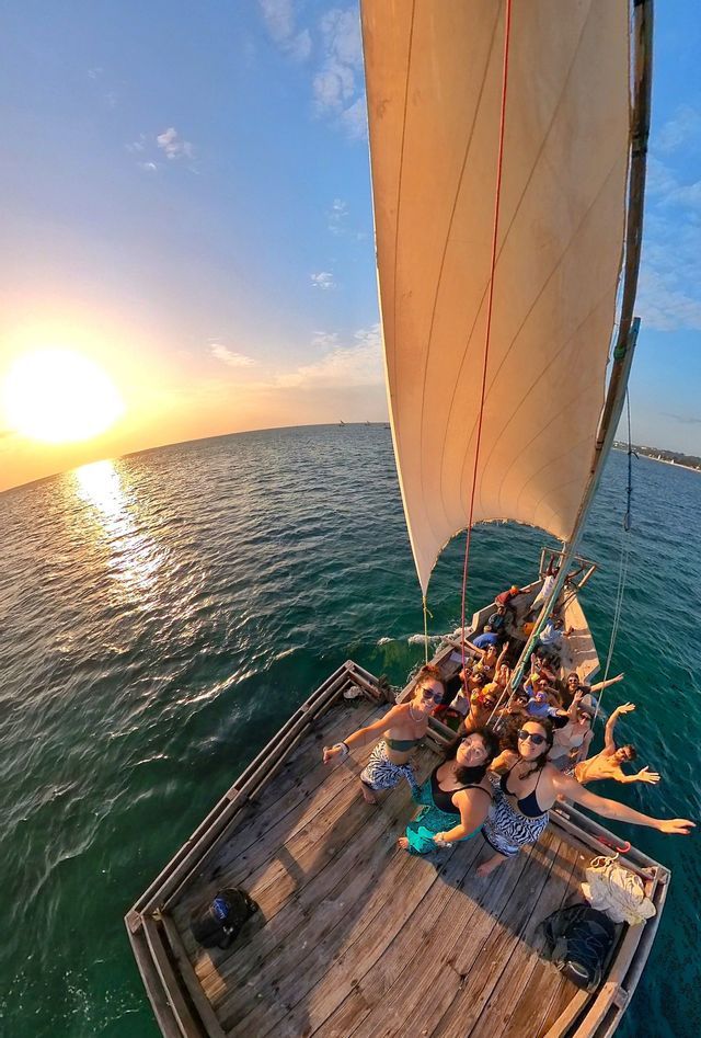 Un groupe WeRoad prend une photo en plongée sur un voilier en bois, naviguant sur l'océan au coucher du soleil.