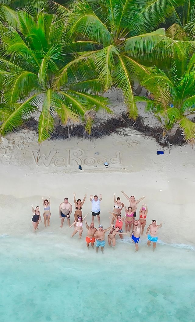 Una vista aérea de un viaje en grupo de WeRoad saludando desde el agua turquesa de una playa de arena blanca con 'WeRoad' escrito en la arena.