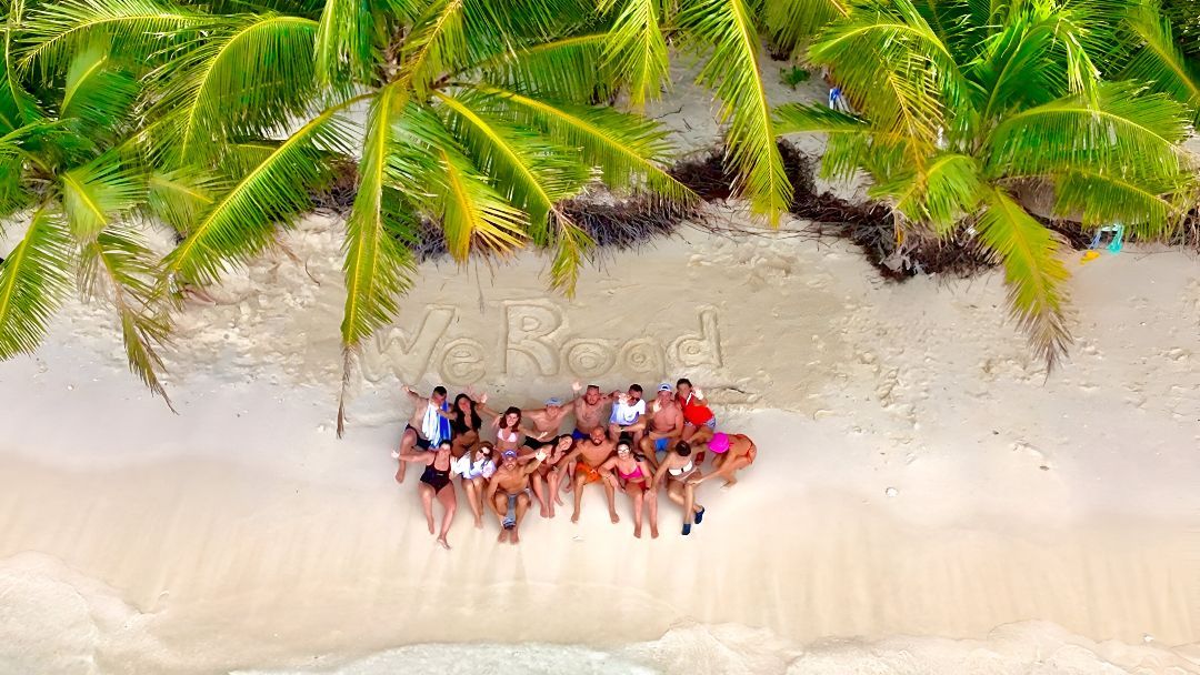 Vista aérea de un viaje en grupo de WeRoad posando en una playa de arena blanca bajo palmeras, con 'WeRoad' escrito en la arena.
