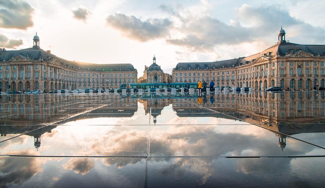 Un gran edificio clásico y un cielo nublado se reflejan en el suelo mojado de una gran plaza pública al atardecer.