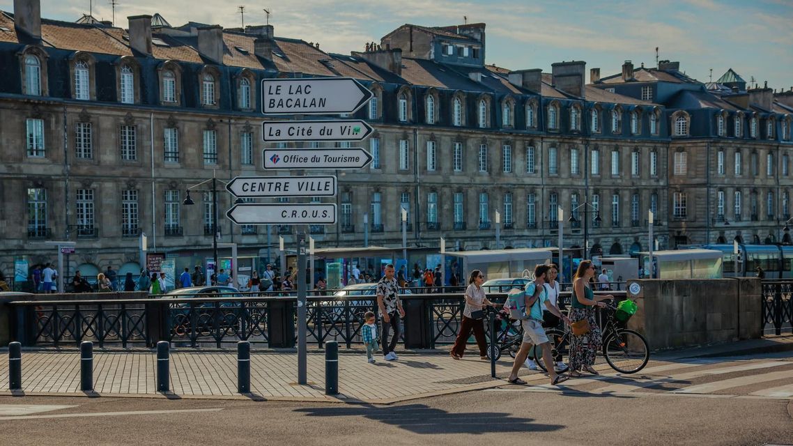 Segnali stradali direzionali bianchi in francese si trovano di fronte a un grande edificio classico, mentre i pedoni attraversano la strada in una giornata di sole.