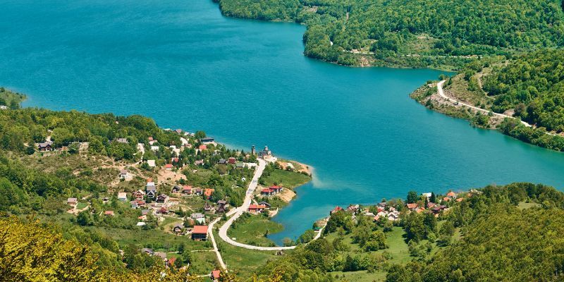Eine Luftaufnahme eines kleinen Dorfes am Ufer eines hellblauen Sees, eingebettet in dichte, grüne, bewaldete Hügel.