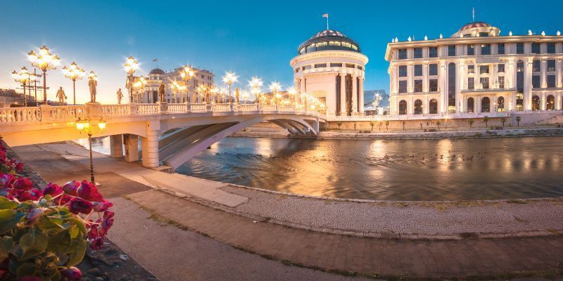 Eine beleuchtete Brücke wölbt sich bei Dämmerung über einen Fluss in einer Stadt, gesäumt von verzierten Gebäuden und Laternen am Ufer.