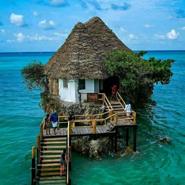 A small, thatched-roof building sits on a rock in the turquoise ocean, with people on a wooden walkway leading to it.