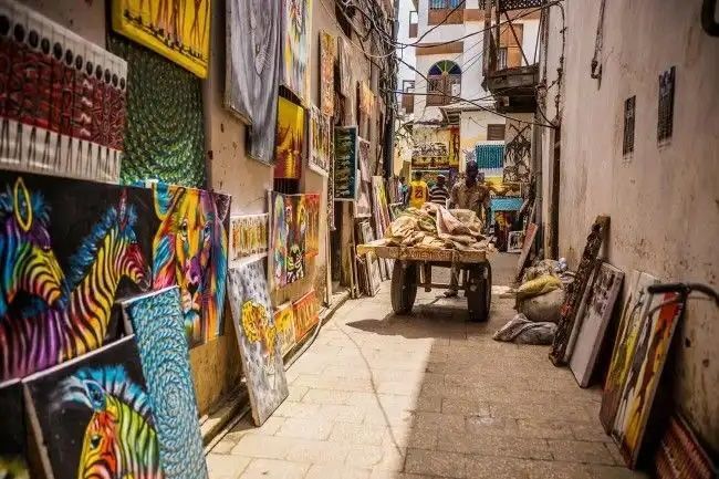 Colorful paintings of animals and patterns are displayed for sale along the walls of a narrow sunlit alley.