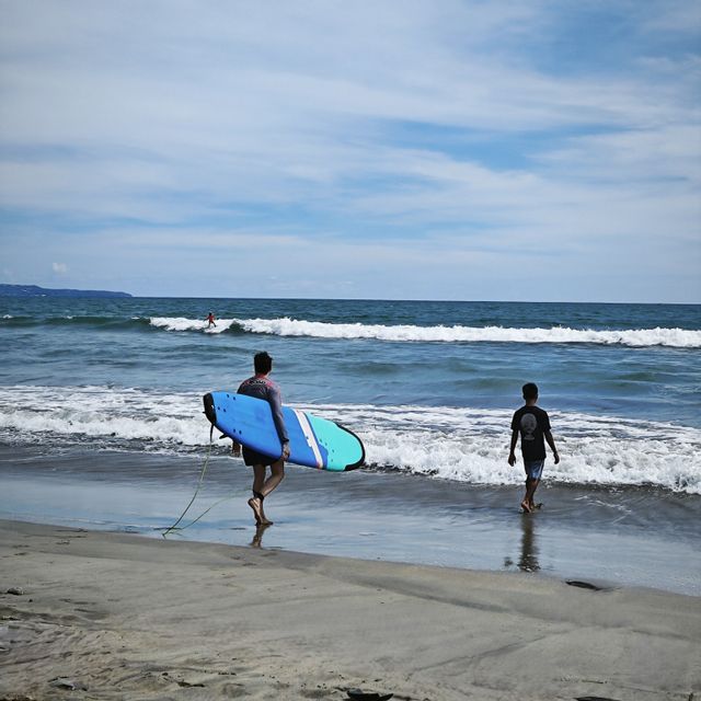Eine Person mit blauem Surfbrett und ein Junge gehen am Strand entlang in die Meereswellen unter einem teilweise bewölkten Himmel.