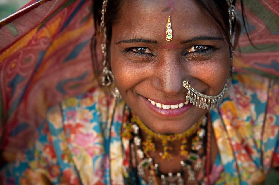 Primo piano di una donna sorridente che indossa abiti tradizionali colorati, con un anello al naso decorativo e un bindi sulla fronte.