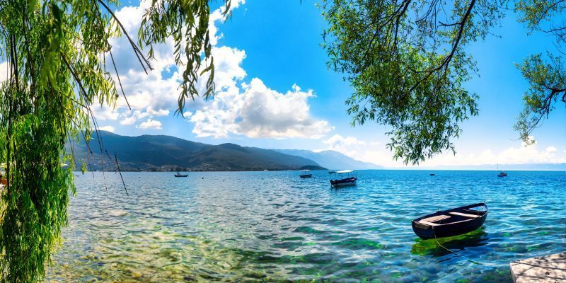 Ein kleines Boot liegt an einem klaren, ruhigen See mit entfernten Bergen, umrahmt von grünen Ästen unter blauem Himmel.
