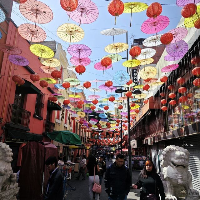 Bunte Papierschirme und rote Laternen hängen über einer belebten Stadtstraße, wo Menschen zwischen Gebäuden spazieren.
