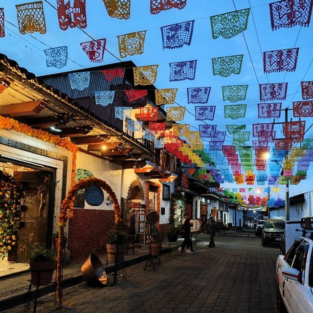 Bunte Papel Picado Banner hängen in der Abenddämmerung über einer engen Kopfsteinpflastergasse, die von mit Ringelblumengirlanden geschmückten Gebäuden gesäumt ist.