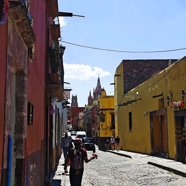 Eine Person geht eine schmale Kopfsteinpflastergasse entlang, die von bunten Häusern gesäumt ist, mit Kirchtürmen, die in der Ferne unter blauem Himmel sichtbar sind.